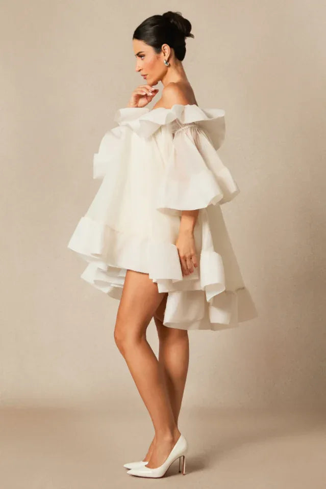 Model wearing a white off-the-shoulder dress with voluminous ruffles and bell sleeves, standing in profile with one hand touching her chin.