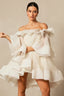 Model wearing a white off-the-shoulder dress with voluminous ruffles and bell sleeves, posing with arms crossed.
