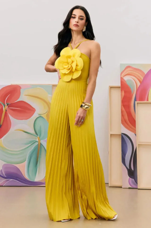 Side view of a woman in a yellow pleated jumpsuit with a floral bust detail, posing with hand on hip. Secondary image