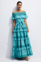 Woman wearing a turquoise off-the-shoulder maxi dress with tiered ruffles and lace trim, standing against a white background.