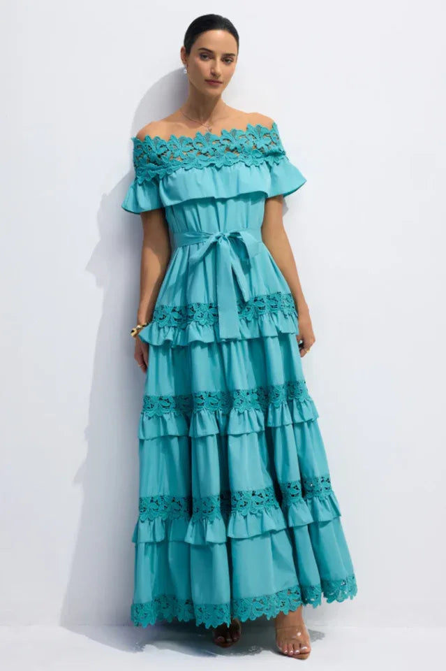 Woman wearing a turquoise off-the-shoulder maxi dress with tiered ruffles and lace trim, standing against a white background.