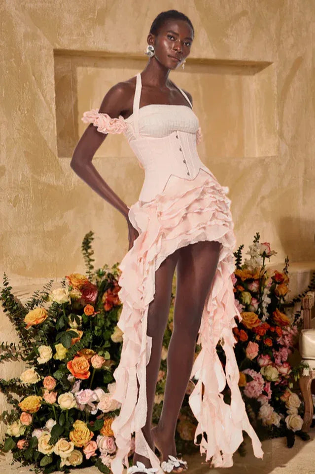 Model wearing a pink ruffled corset dress with a halter neckline and high-low hemline, standing in front of a floral arrangement. Main image