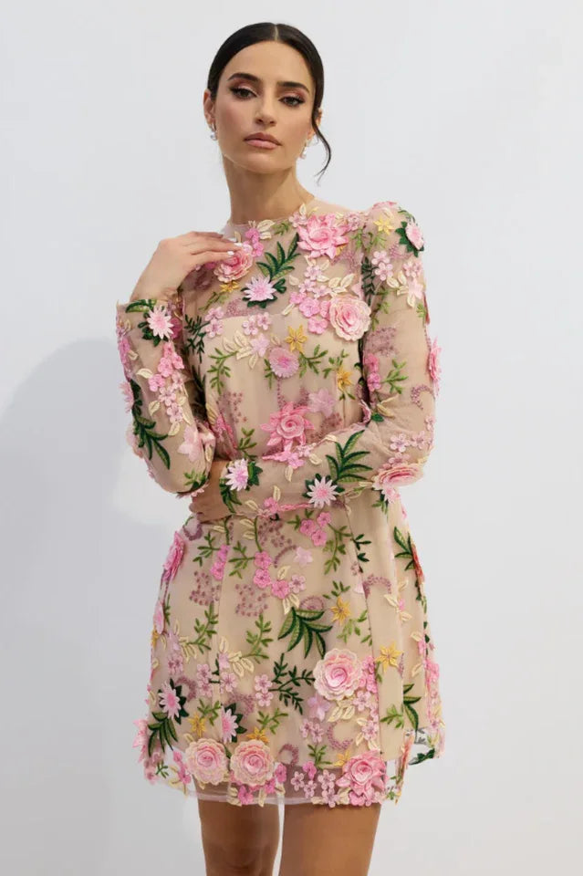 Model wearing a short, long-sleeved dress with a nude base and 3D floral embroidery in pink, yellow, and green.
