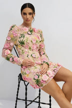 Model sitting on a chair wearing a sheer, floral-embroidered dress with long sleeves.