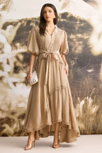Woman wearing a tan linen blend maxi dress with puffed sleeves and a tiered skirt, holding a shell-shaped clutch.