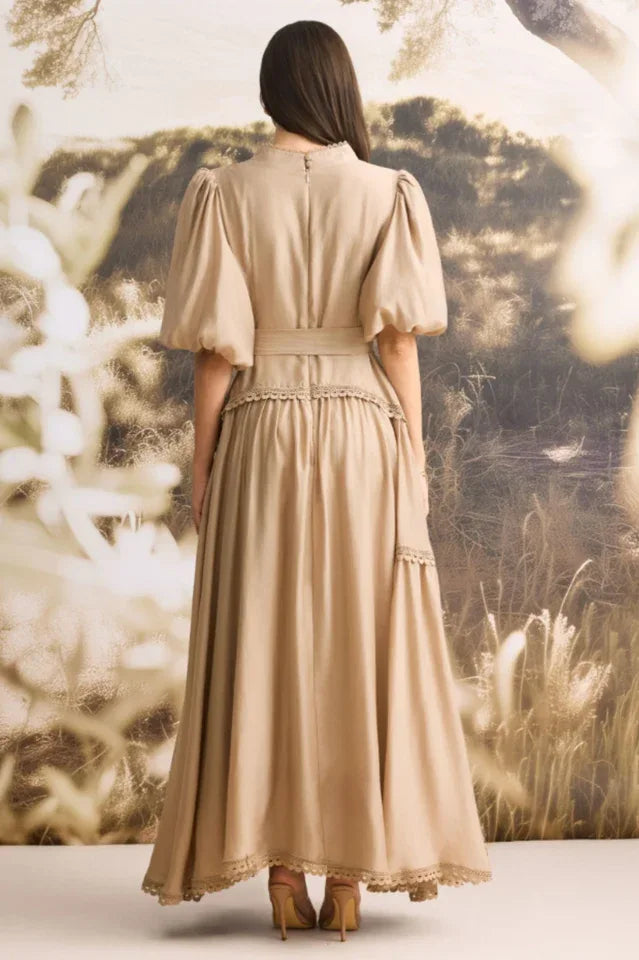 Back view of a woman wearing a tan linen blend maxi dress with puffed sleeves and a tiered skirt.