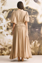 Back view of a woman wearing a tan linen blend maxi dress with puffed sleeves and a tiered skirt.