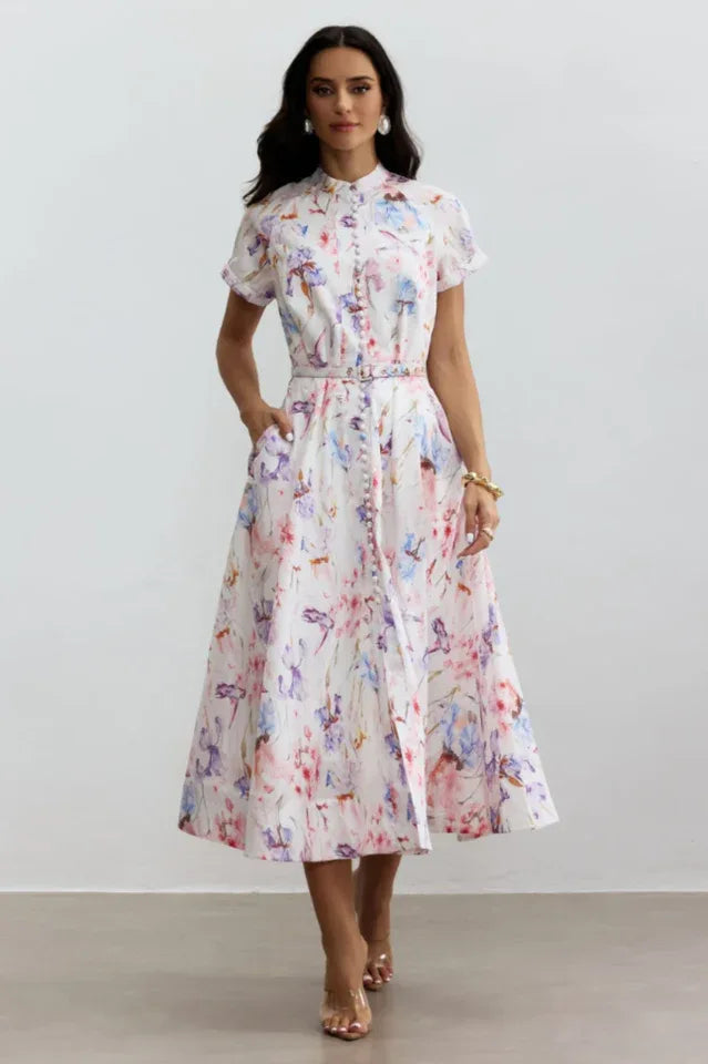 Woman wearing a white midi dress with a floral print, short sleeves, and a belted waist, standing against a white background.