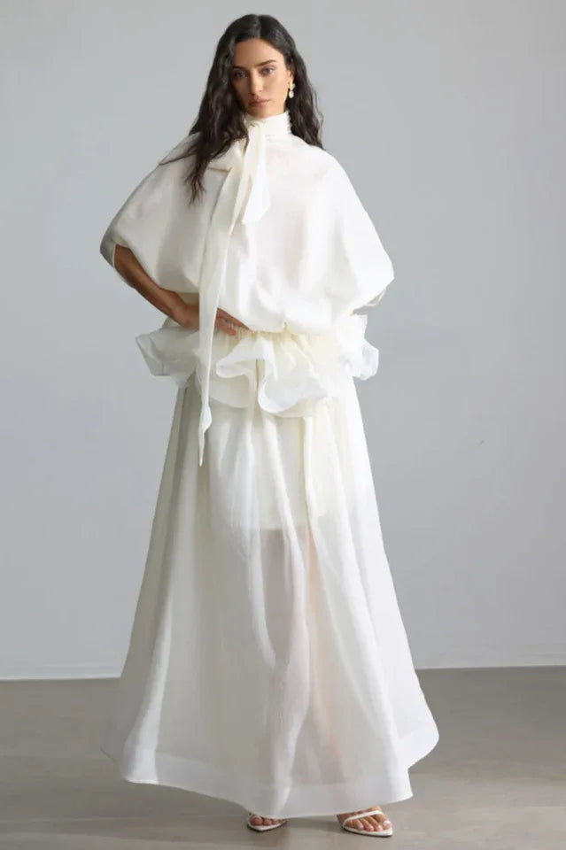Woman wearing a white, flowing, high-neck dress with voluminous sleeves and a tiered ruffled waist. Main image