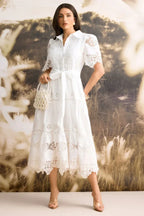 Woman wearing a white collared midi dress with lace detailing and a belted waist, holding a beaded handbag.
