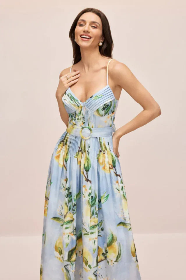 Woman in a light blue midi dress with a lemon print, smiling and touching her chest.