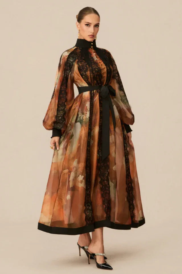 Full-body shot of a model in an orange and brown printed maxi dress with a black belt and high neckline. Secondary image