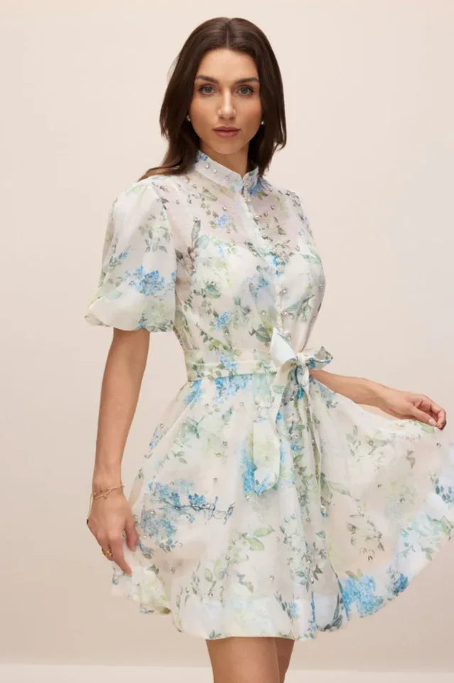 Woman looking at the camera wearing a white floral print mini dress with puff sleeves and a belted waist Secondary image