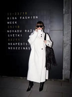 Woman in a white oversized wool blend coat standing in front of a black wall with event details