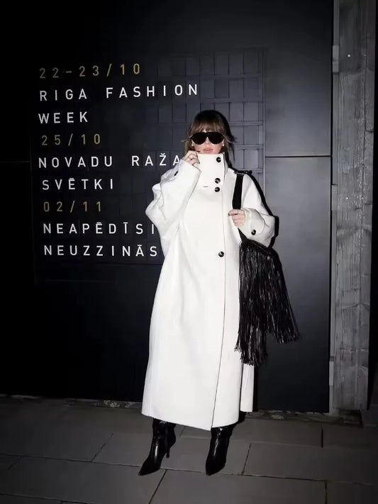 Woman in a white oversized wool blend coat standing in front of a black wall with event details