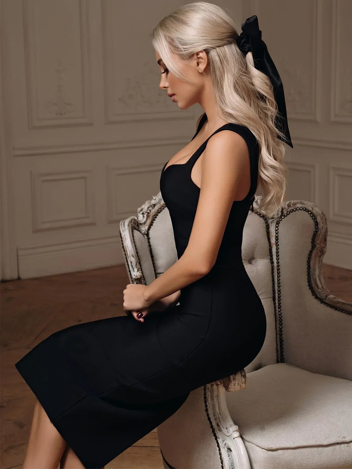 Side view of a woman in a black sleeveless knee-length dress with a plunging v-neck, sitting on a chair indoors.