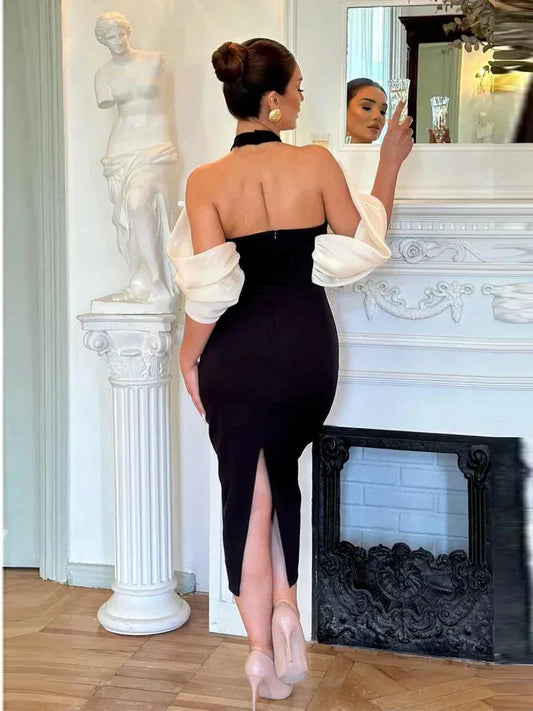 Back view of a woman in a black sheath dress with a white sheer off-the-shoulder overlay and flower accent, holding a champagne flute.