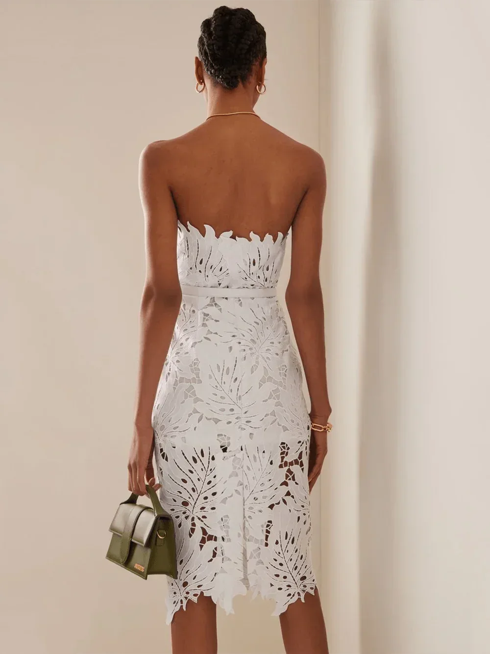 Rear view of a woman in a white strapless lace sheath dress with leaf cutouts and a green handbag.