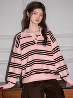 Woman wearing a pink and black striped knit polo sweater and brown pants, leaning on a wooden surface.