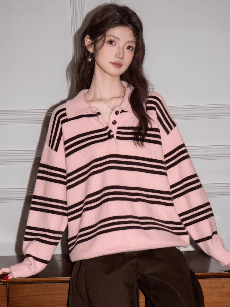 Woman wearing a pink and black striped knit polo sweater and brown pants, leaning on a wooden surface.