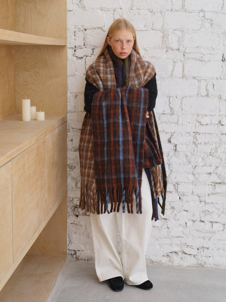 A young woman with blonde hair and freckles stands holding a large, plaid scarf. The scarf has a pattern of brown, blue, and pink squares.