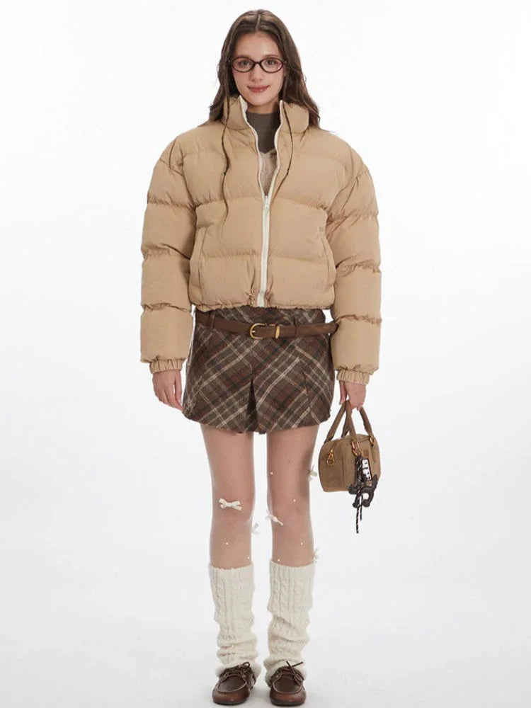 Model wearing a tan cropped puffer jacket, brown plaid pleated mini skirt, white leg warmers, and brown loafers.
