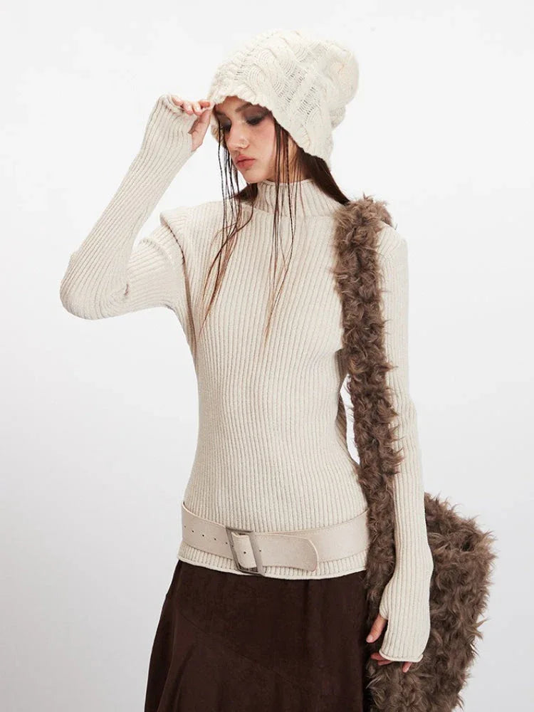Close-up of a model wearing a cream ribbed turtleneck sweater, cream belt, and a cream knit beanie, touching her beanie.