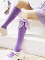 Model wearing purple knee-high socks with large decorative bows on the side of each leg, paired with white platform Mary Jane shoes.