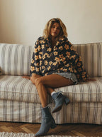 Woman sitting on a striped couch, wearing a grey fleece jacket with a yellow floral print and denim shorts.