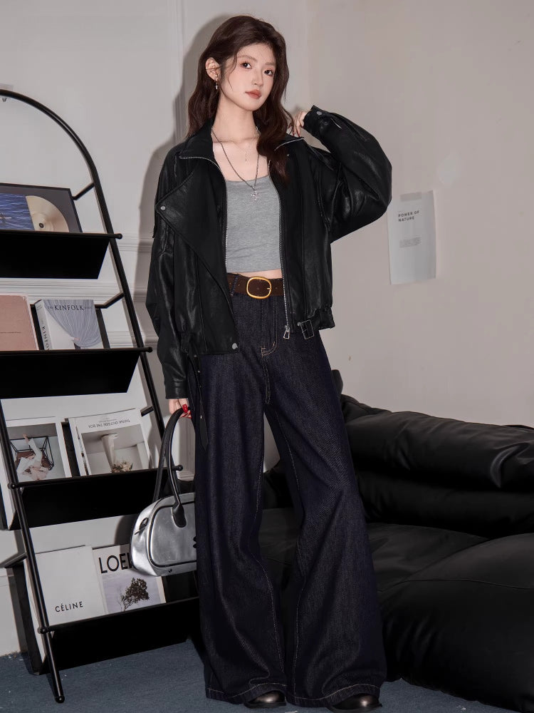 Full body shot of a woman in a black faux leather jacket, grey crop top, dark wash flared jeans, and carrying a silver handbag.