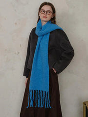Woman wearing a dark grey sweater and a bright blue textured knit scarf with fringe, standing against a textured wall.