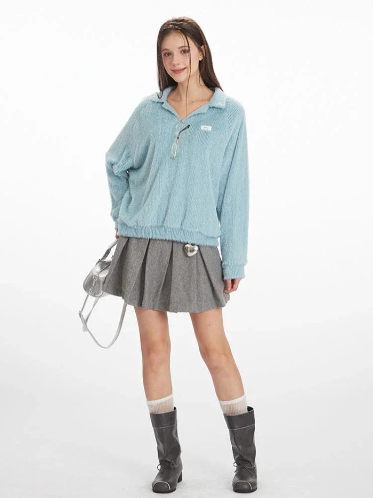 Full body shot of a model wearing a light blue fuzzy knit collared v-neck sweater with a grey pleated mini skirt, white and grey striped socks, and black boots, holding a silver handbag.