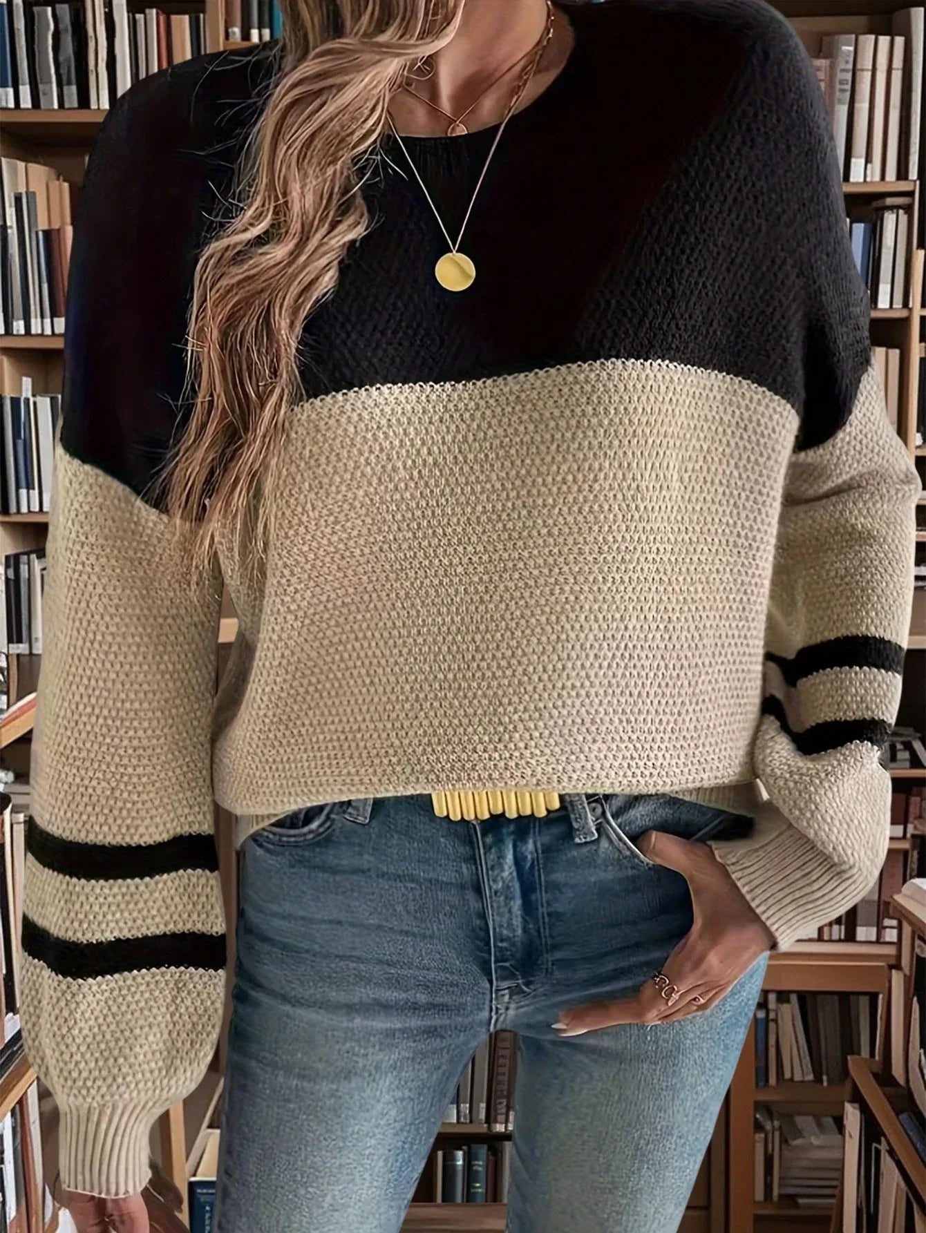 Woman modeling a color-blocked sweater with a textured knit pattern and striped sleeves, paired with blue jeans.