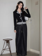 Woman standing in a fitting room wearing a black and grey layered top with ruffled trim and dark wide-leg jeans, holding a silver handbag.