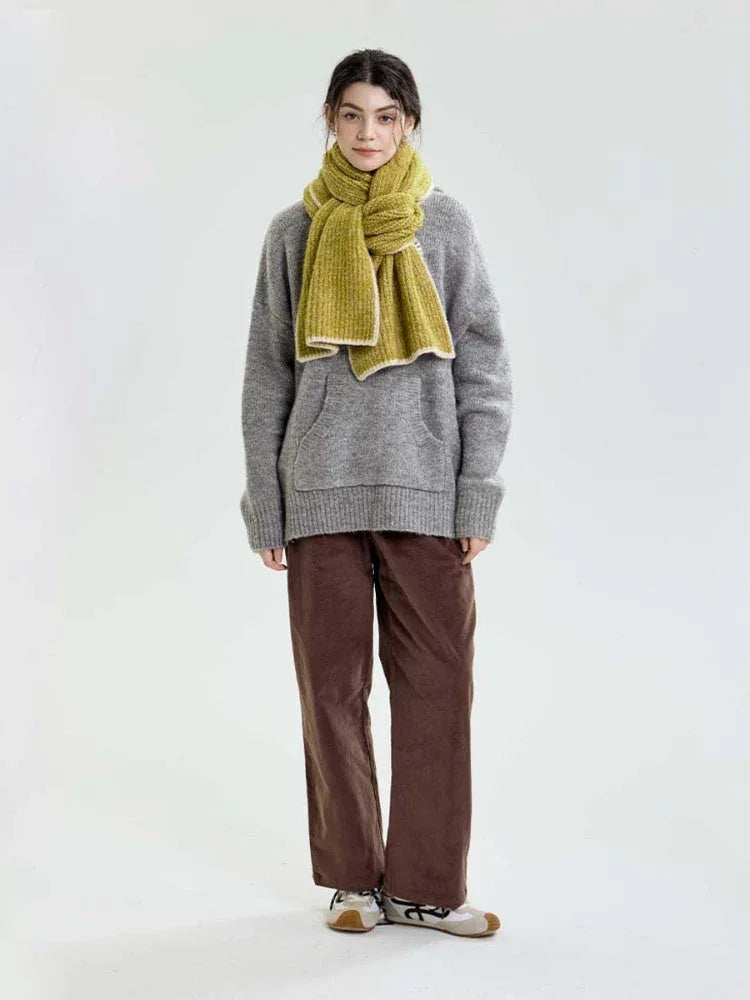 Full-length view of a model in a grey sweater and brown pants, accessorized with a lime green chunky knit scarf.