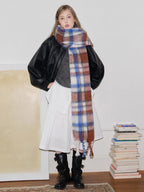 Full body shot of a model in a black jacket, grey sweater, white skirt, and a large plaid scarf.