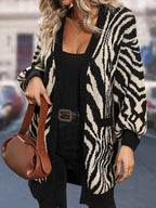 Woman wearing a black and white zebra print longline knit cardigan over a black top and black pants, holding a brown handbag.