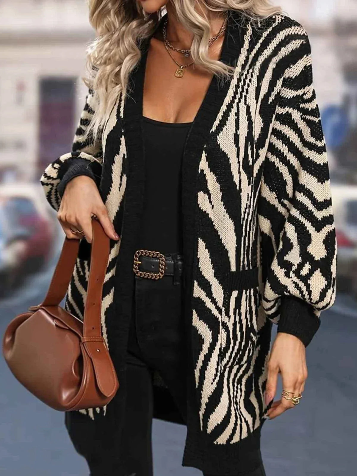 Woman wearing a black and white zebra print longline knit cardigan over a black top and black pants, holding a brown handbag.