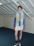 Full body shot of a woman wearing an oversized ombre scarf, grey sweater, blue plaid shirt, and white shorts.