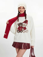 Model adjusting a red scarf worn with a white fuzzy knit sweater featuring a teddy bear graphic.