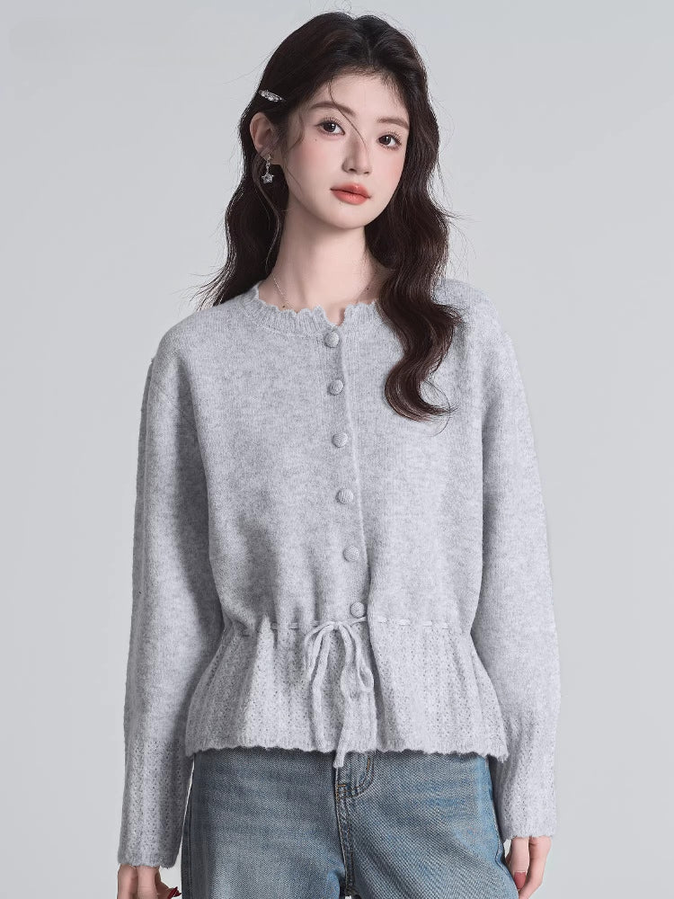 A woman stands wearing a light grey knit cardigan with a drawstring waist and button front closure, paired with blue jeans.