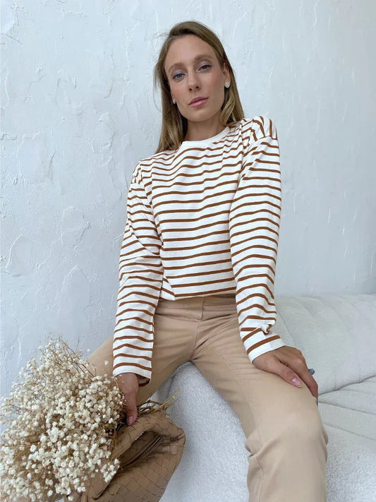 Woman wearing a white and brown striped long-sleeve top and beige pants, sitting on a couch with a bouquet of dried flowers and a beige handbag.