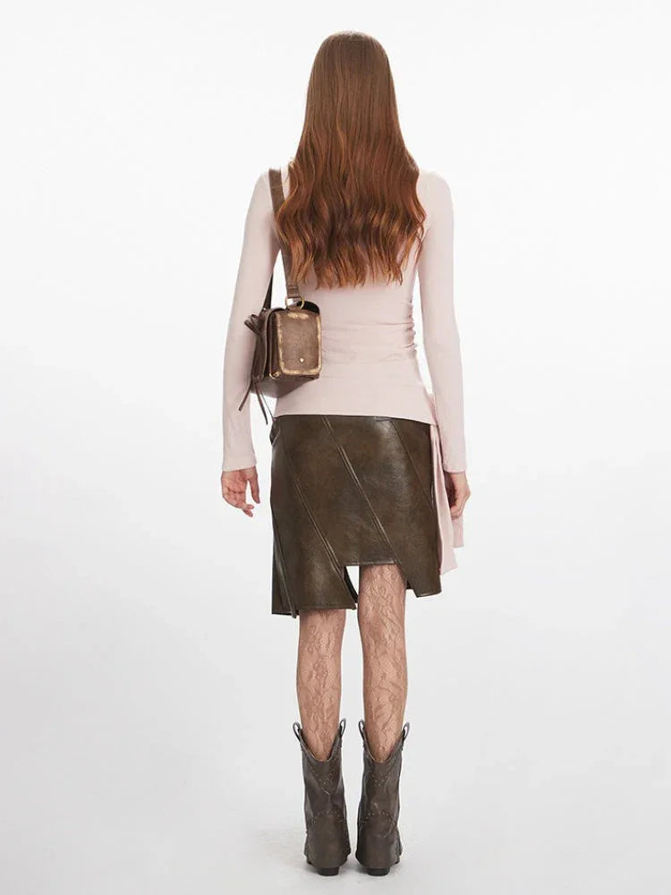 Full-body back view of a woman wearing a pink long-sleeve ruched top, a brown asymmetrical faux leather skirt, patterned tights, and dark boots.