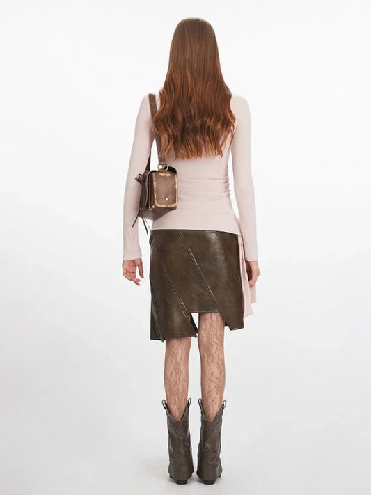 Full-body back view of a woman wearing a pink long-sleeve ruched top, a brown asymmetrical faux leather skirt, patterned tights, and dark boots.