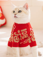 A white cat wearing a red knit sweater with gold Chinese character designs, looking to the left.