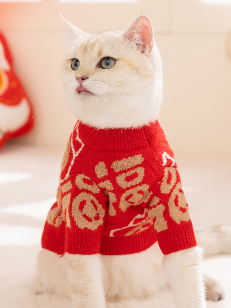 A white cat wearing a red knit sweater with gold Chinese character designs, looking to the left.