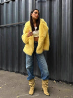 Woman in a yellow faux fur jacket over a white crop top and blue jeans, with beige boots, standing against a corrugated metal wall.