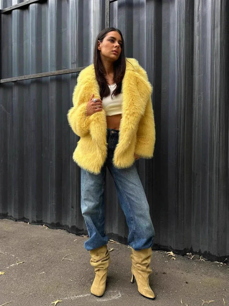 Woman in a yellow faux fur jacket over a white crop top and blue jeans, with beige boots, standing against a corrugated metal wall.