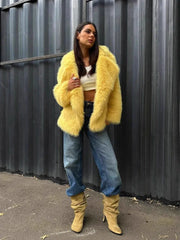 Woman in a yellow faux fur jacket over a white crop top and blue jeans, with beige boots, standing against a corrugated metal wall.