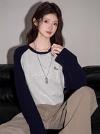 Woman sitting on a black couch, wearing a light grey long-sleeved t-shirt with navy blue raglan sleeves and beige pants.